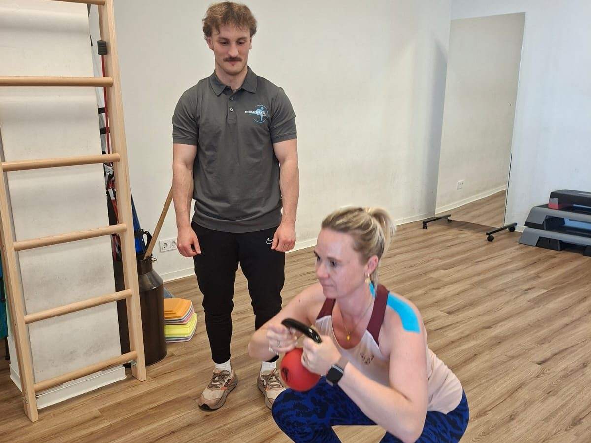 Personal Training im Therapiepark Augsburg: Frau macht eine Kniebeuge mit Kettlebell, Mann im Praxisshirt schaut zu