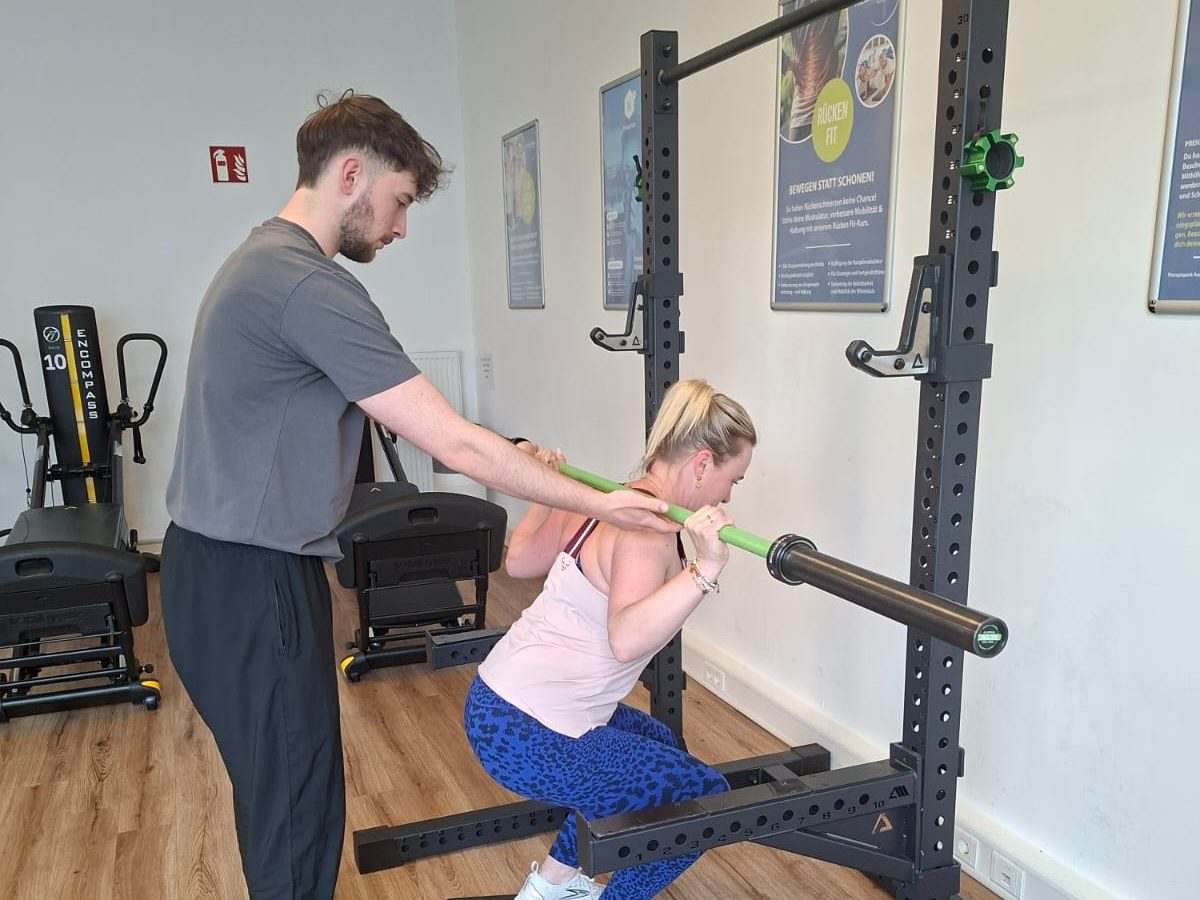 Training im Therapiepark Augsburg: Frau macht eine Kniebeuge mit Stange, ein Mann im grauen Shirt greift an die Stange